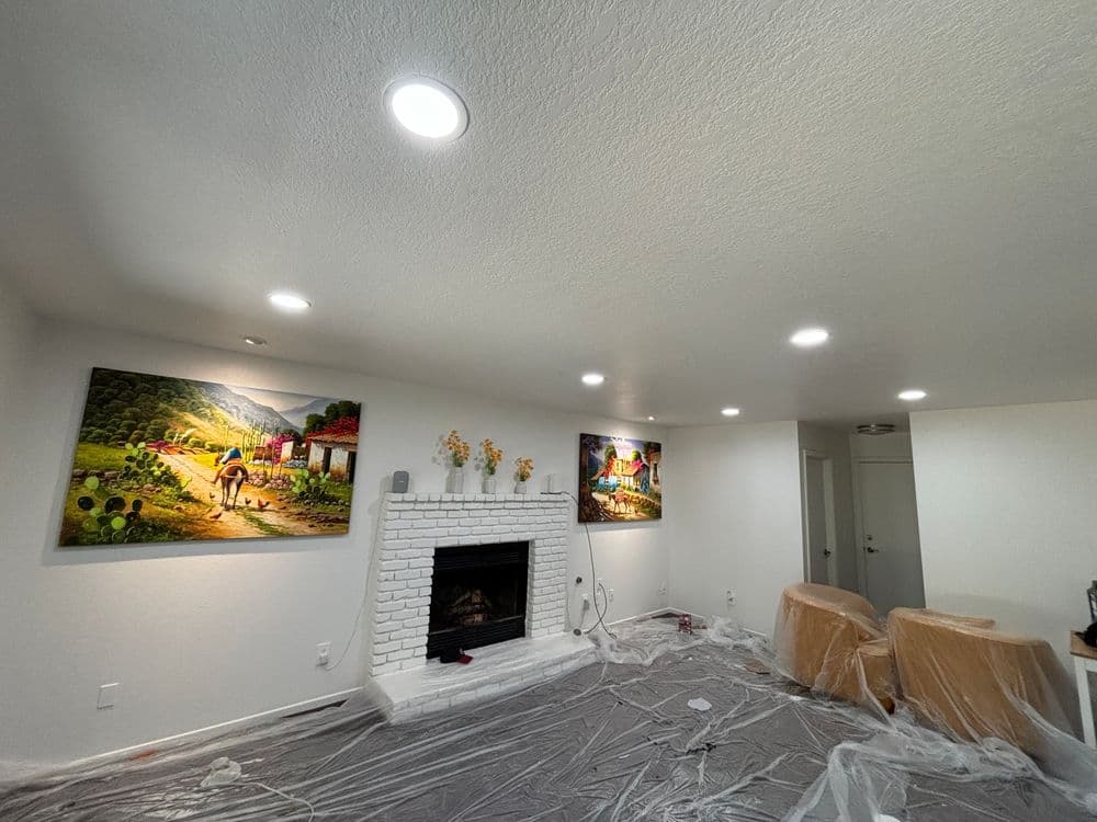 Living room with white walls, bright recessed lights, and paintings, featuring a fireplace and plastic cover.