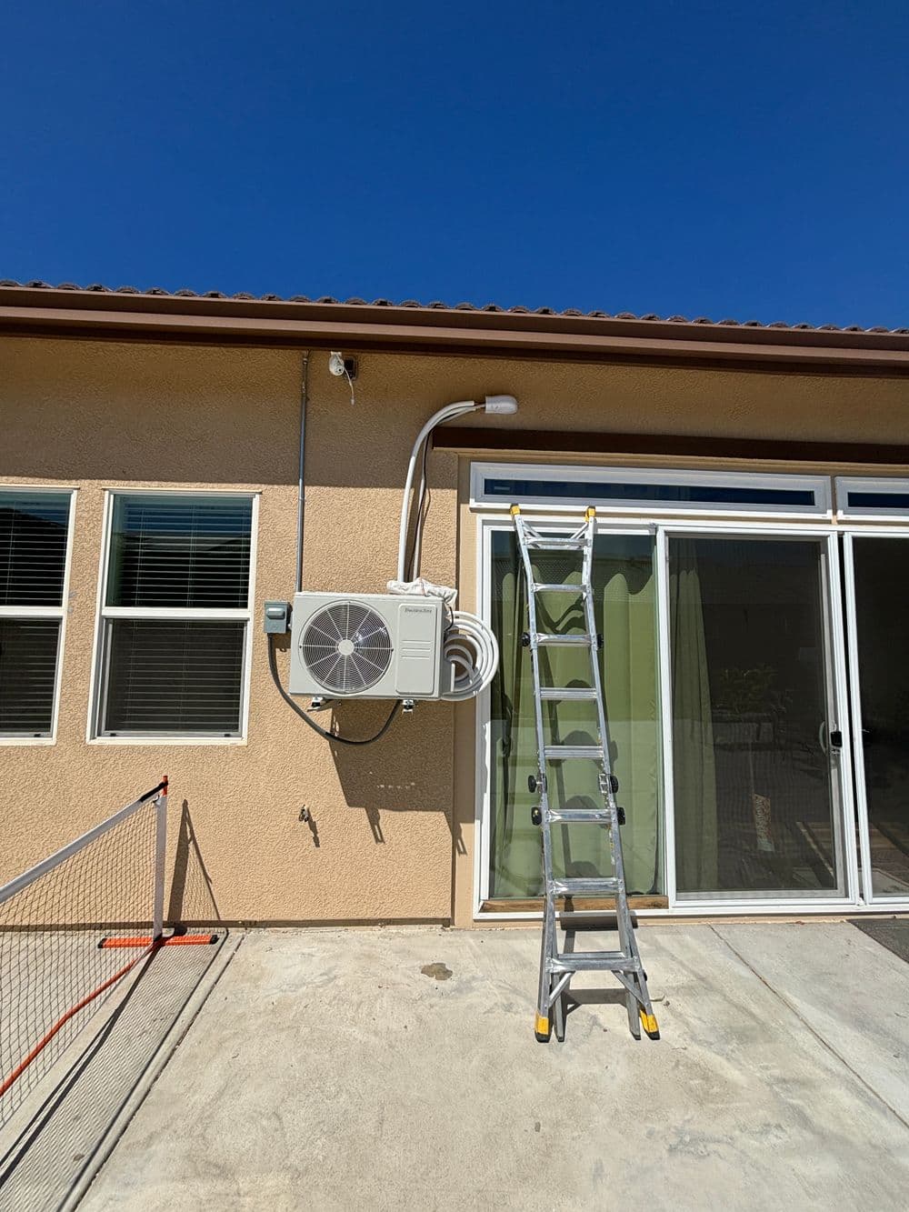 Air conditioning unit mounted on exterior wall with extension ladder nearby, blue sky backdrop.