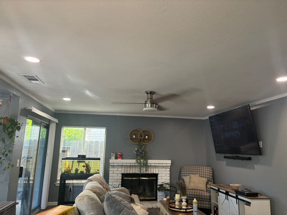 Modern living room with ceiling fan, TV, white brick fireplace, and natural light.