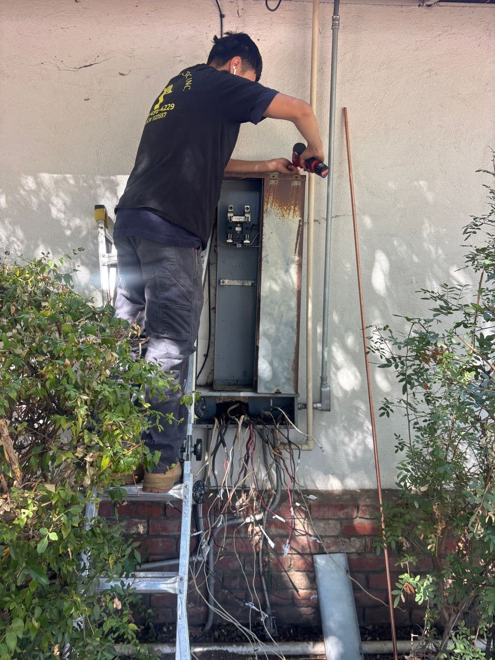 Technician using a drill on an electrical panel while standing on a ladder outdoors.